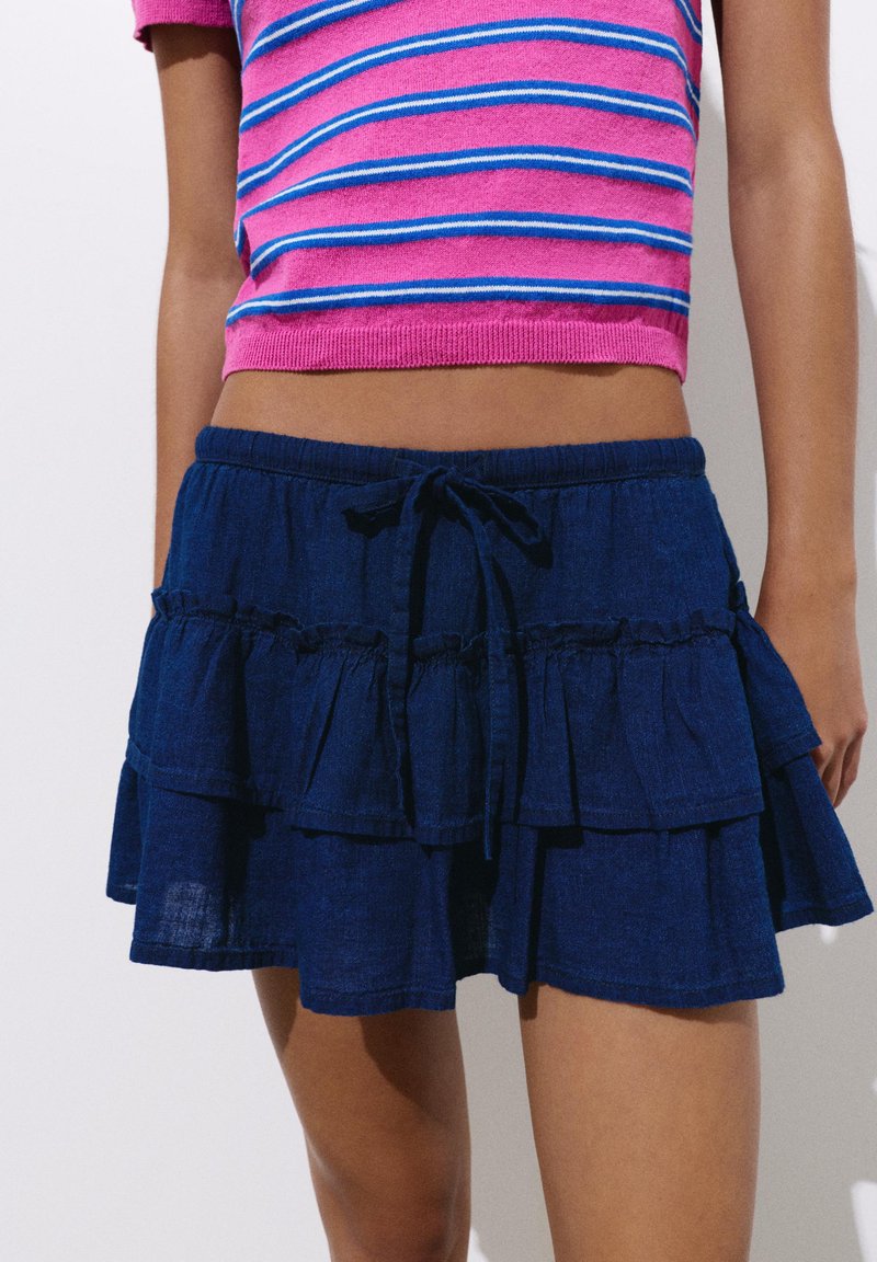 Personne portant une mini-jupe bleue à volants avec une taille à cordon et un crop top rose à rayures manches courtes, debout sur un fond blanc.