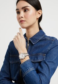 Orologio argento con quadrante rotondo e tre subdiali su un bracciale metallico, indossato su un polso con una giacca di denim con zip e colletto.