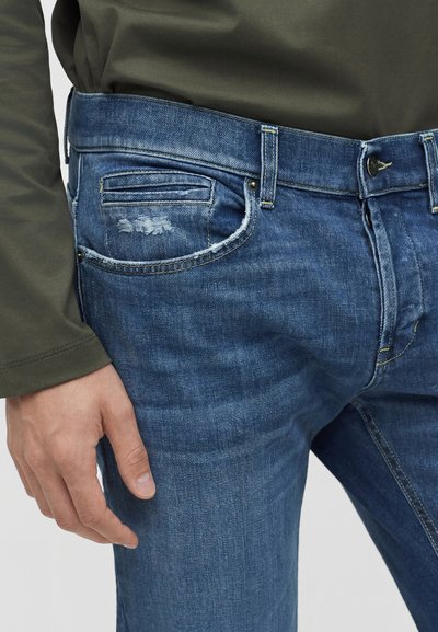 Gros plan d'un jean en denim bleu avec une légère usure près de la poche avant, porté avec une chemise longue manche de couleur olive.