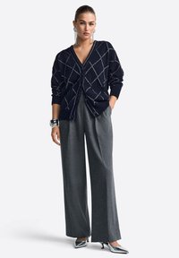 Tumesinine kardigan, millel on teemantmuster, nupud ja taskud, kombineeritud laiade hallide pükste ja metallik kõrge kontsaga kingadega.