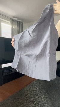 Persona che tiene un vestito senza maniche, grigio chiaro e spiegazzato, in un soggiorno con una TV e delle tende sullo sfondo.