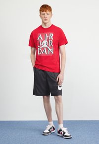Red cotton t-shirt with white graphic "AIR JORDAN" design, paired with black shorts featuring a white Nike logo and black-and-white sneakers.