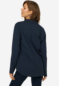 Camisa de manga larga de color azul marino con cuello, corte relajado, detalle en el yugo trasero y dobladillo redondeado. La tela parece suave con una sutil textura.