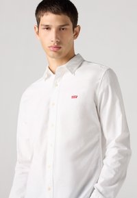 Baltas medvilninis marškinėlių su kietu audiniu. Turi klasikinę apykaklę ir mažą raudoną Levi's logotipą kairiajame krūtinės gale.