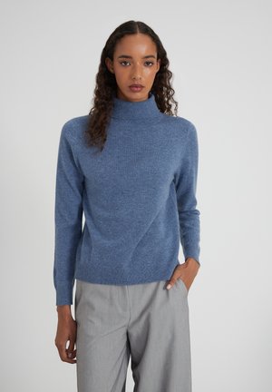 Femme aux cheveux bouclés portant un pull col roulé bleu et un pantalon gris clair, se tenant avec une main dans la poche devant un fond uni.
