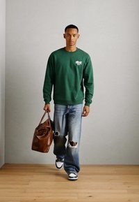 Groene sweater met een klein wit logo, gecombineerd met lichtblauwe ripped jeans en zwart-witte sneakers. Bruin leren tasje in de hand.