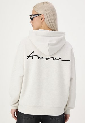 Femme aux longs cheveux blonds portant des lunettes de soleil noires et un sweat à capuche blanc avec "Amour" écrit dans le dos en lettres cursives noires.
