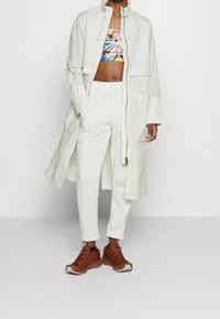 Osoba oblečená v světle béžovém dlouhém kabátu, bílých teplákových kalhotách, barevném květinovém topu a hnědých teniskách, stojí proti jednobarevnému pozadí.