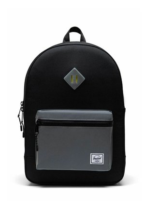 Sac à dos noir avec une grande poche avant zippée grise et un petit patch en forme de diamant au-dessus, avec une poignée de transport en haut.