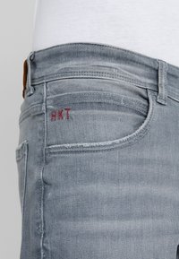 Jeans de mezclilla azul claro con un acabado desgastado. Presenta un logo rojo bordado "HKT" en el bolsillo trasero. Diseño estándar de cinco bolsillos.