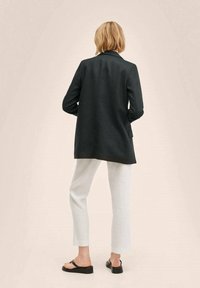 Tmavě šedý blazer se střihem na míru, s jediným rozparkem vzadu, spárovaný se světle šedými krátkými kalhotami a černými plochými sandály. Hladká, tkaná textura.