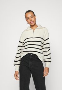 Strikket genser i kremfarge og svarte striper, med en halv glidelås og voluminøse ermer, kombinert med høyt liv svart jeans.