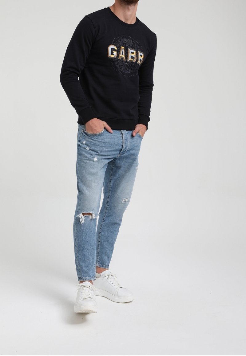 Homme portant un sweat-shirt noir à manches longues avec le texte "GABB", un jean bleu clair déchiré et des baskets blanches, debout les mains dans les poches.