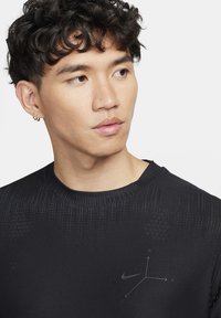 Nuori mies, jolla on kihara tumma hius ja pienet rengaskorvakorut, pukeutuneena mustaan Nike-paitaan, jossa on hienovarainen 3D-koordinaattimuotoilu.