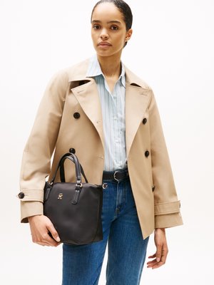 Schwarze Handtasche mit strukturiertem Design, glatter Textur, kurzen Griffen und goldenen Hardware-Akzenten, gehalten von einer Person, die einen beigen Trenchcoat und ein blaues Hemd trägt.