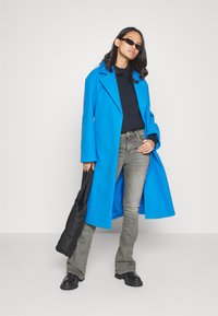Cappotto lungo blu con ampi revers, indossato sopra un maglione nero. Jeans grigi a zampa d'elefante e stivali neri chunk. Borsa nera a tracolla.