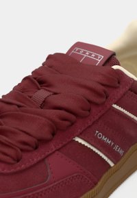 Červená teniska s vrchom zo semišu a textilu, hrubými šnúrkami a bielymi pruhmi. Logo viditeľné na jazyku a boku. Gumená podrážka.