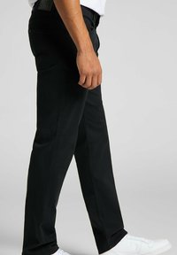 Pantalones negros de corte ajustado, con una textura suave, costuras visibles y bolsillos laterales. Llevados con zapatillas blancas.