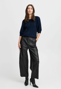 Marineblauer gestrickter Pullover mit einem strukturierten Rautenmuster, kombiniert mit weit geschnittenen schwarzen Lederhosen und spitzen schwarzen High-Heels.