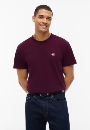 Lachende man staat met handen achter zijn rug, draagt een kastanjebruin Tommy Jeans T-shirt en donkerblauwe spijkerbroek met een zwarte riem.