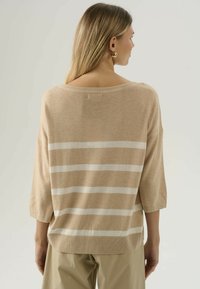 Maglione beige a lavorazione a maglia con strisce orizzontali bianche, scollo ampio, maniche a 3/4 e una vestibilità morbida. Il tessuto appare soffice e ha una texture liscia.