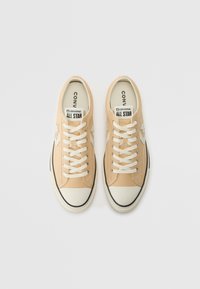 Bež platnene teniske s bijelim naglascima, okrugli vrh, kontrastna crna guma potplat i središnji sustav vezanja. Sadrži "Converse All Star" brendiranje.