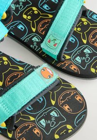 Svarta sandaler med turkosa remmar som har Pokémon-figurer i olika färger. Mjukt material med justerbara stängningar för att passa.