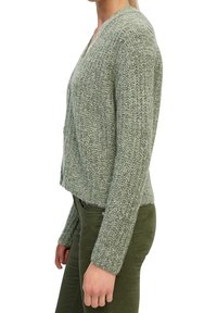 Hellgrüne Strickjacke mit V-Ausschnitt, langen Ärmeln und strukturiertem Muster, kombiniert mit olivgrünen Hosen.