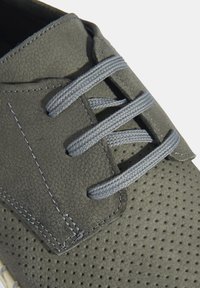 Chaussure en cuir perforé gris avec des lacets gris, au design minimaliste et à la semelle blanche contrastante. Présente une texture lisse et des coutures soignées.