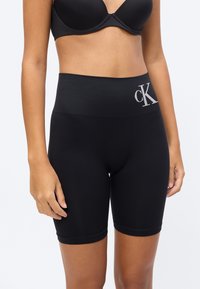 Γυναίκα που φοράει μαύρο ποδηλατικό σορτς ψηλής μέσης με λογότυπο "cK" και μαύρο σουτιέν, σε απλό φωτεινό φόντο.