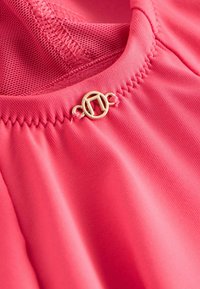 Tissu rose à texture lisse avec une doublure en filet ; présente un fermoir circulaire doré avec un logo angulaire près de l'encolure.