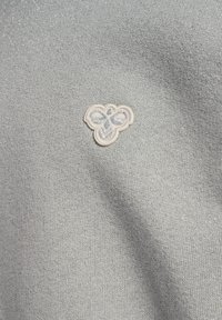 Lys grå, tekstureret stof med en subtil glans; har et broderet bi-logo i lys beige og blå accenter på brystområdet.