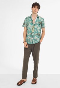 Camisa de manga corta en color verde azulado con patrones de hojas blancas y verdes, combinada con pantalones marrones a rayas y sandalias marrones tipo slides. Los accesorios incluyen gafas de sol amarillas.