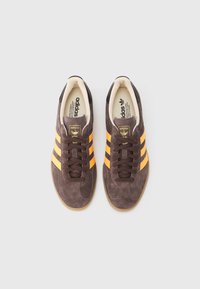 Smeđe tenisice od brušene kože sa žutim i crnim prugama, okruglim vrhom, vezicama za vezanje i potplatom od gume boje gume. Brendirano na jeziku.