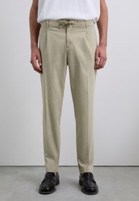 Pantalones beige hechos de una tela ligera; cuentan con una cintura con cordón, dos bolsillos frontales y una silueta ajustada. Llevados con zapatos negros.