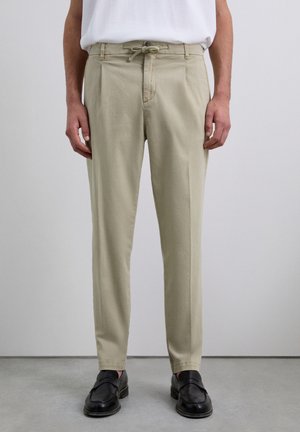 Pantalones beige hechos de una tela ligera; cuentan con una cintura con cordón, dos bolsillos frontales y una silueta ajustada. Llevados con zapatos negros.