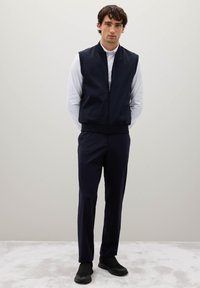 Marineblaues, mit Reißverschluss versehenes Vest über einem weißen Langarmshirt, kombiniert mit passenden marineblauen Hosen und schwarzen Sportschuhen. Glatte, elegante Stoffoberfläche.