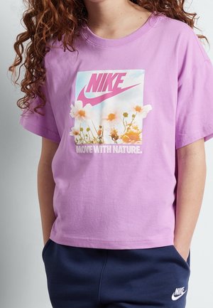Persona que lleva una camiseta morada de Nike con un diseño floral y el texto "MOVE WITH NATURE", combinada con pantalones azul marino de Nike.