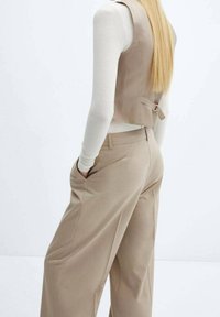 Beżowa kamizelka o dopasowanym kroju z tylnym wiązaniem, zestawiona z szerokimi beżowymi spodniami. Stylizacja zawiera dopasowaną białą bluzkę z długim rękawem.