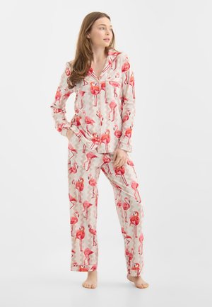 Vrouw die blootsvoets staat, gekleed in crèmekleurige pyjama met een roze flamingopatroon, handen in de zakken, en naar de zijkant kijkt.