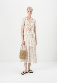 Robe légère à motif floral en crème avec des manches courtes à volants et un nouage à l'avant. Associée à un sac en osier et des tongs.
