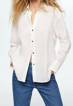 Femme portant une chemise blanche boutonnée ample et un jean bleu, mains dans les poches, avec des cheveux blonds ondulés visibles aux épaules.