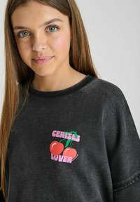 Sweatshirt noir à manches courtes, avec un col côtelé, orné d'un design graphique rose de cerises et du texte "AMATEUR DE CERISES".