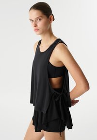 Top negro sin mangas con un diseño fluido, que presenta un detalle de lazo en el costado, espada cortada en la parte posterior y una textura suave. Silueta simple y moderna.