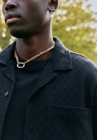 Veste noire texturée avec un motif en losanges, portée sur un haut noir. Les colliers comprennent un cordon tressé et un détail métallique.