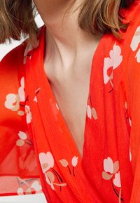 Haut cache-cœur rouge à motif floral en tissu léger. Présente un décolleté en V profond et des manches larges avec des accents de fleurs blanches et roses.