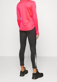 Felpa sportiva a maniche lunghe di un vivace rosa, con schiena texturizzata, abbinata a leggings neri con dettagli a strisce bianche e scarpe nere leggere.