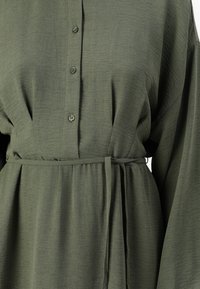 Robe verte olive à boutons avec un tissu texturé, des manches longues, une taille cintrée avec une ceinture à nouer, et une jupe fluide.