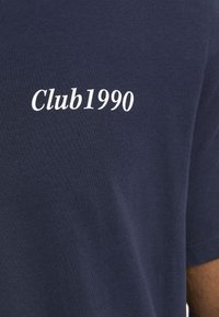 Navyblaue Baumwoll-T-Shirt mit weißem Text "Club1990", der auf der Vorderseite gedruckt ist. Glatte Textur, kurze Ärmeln und schlichtes Design.