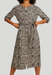 Femme portant une robe léopard mi-longue avec des boutons et des manches trois quarts, mains posées sur les hanches, sur un fond uni.